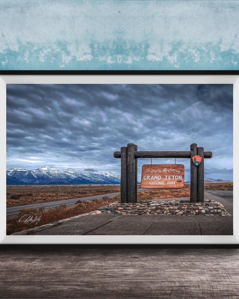 A framed photograph of a sign for Grand Teton National Park, with a cloudy sky and mountains in the background.