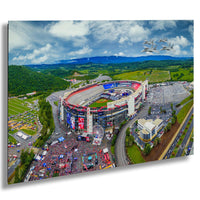 An aerial view of a large sports stadium surrounded by a lush green landscape, with a ferris wheel and other attractions visible. Several planes can be seen flying in the sky above the stadium.