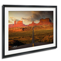 a long, straight road cutting through a vast, arid landscape with towering red rock formations in the background.