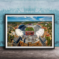 The image is an aerial view of a large sports stadium with a football field, surrounded by buildings and a blue sky.
