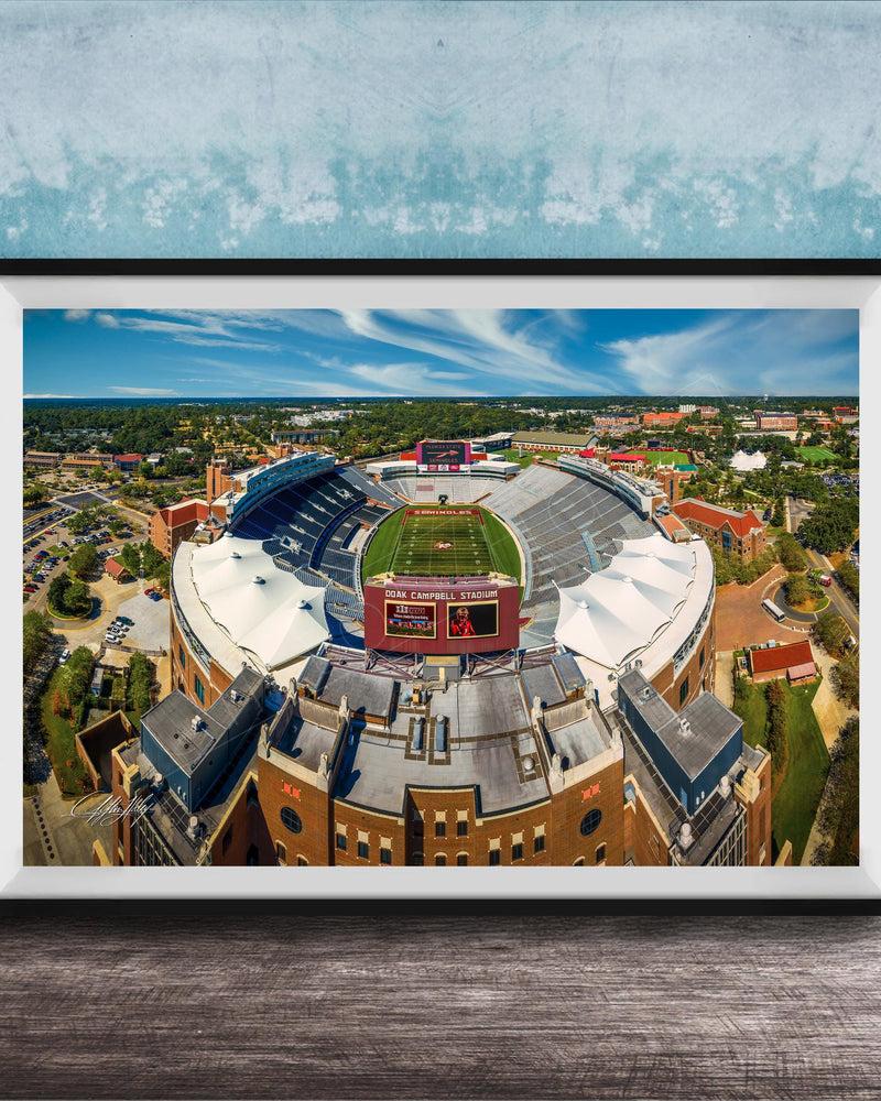The image is an aerial view of a large sports stadium with a football field, surrounded by buildings and a blue sky.