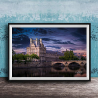 a large, ornate building with a tower and multiple spires, situated on the banks of a river. The sky is filled with clouds, creating a dramatic backdrop for the scene. The building is surrounded by trees, and a bridge can be seen in the background. The image is framed and displayed on a wooden surface, with a blue wall in the background.