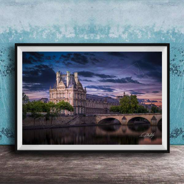 a large, ornate building with a tower and multiple spires, situated on the banks of a river. The sky is filled with clouds, creating a dramatic backdrop for the scene. The building is surrounded by trees, and a bridge can be seen in the background. The image is framed and displayed on a wooden surface, with a blue wall in the background.