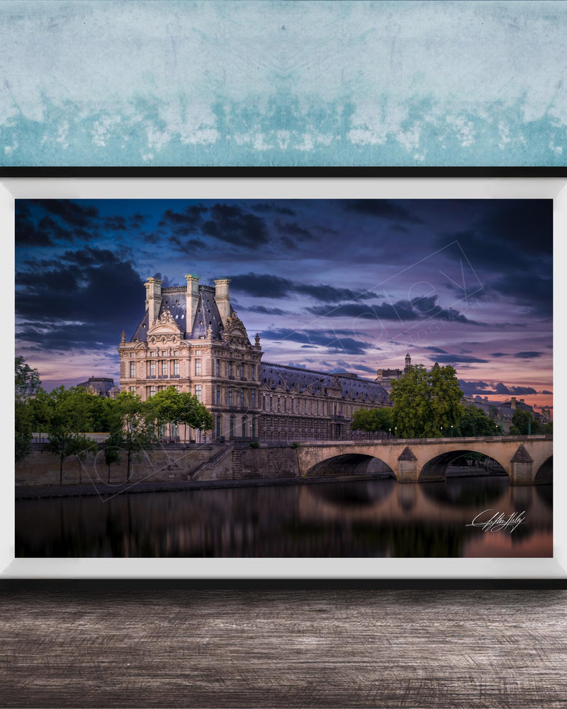 a large, ornate building with a tower and multiple spires, situated on the banks of a river. The sky is filled with clouds, creating a dramatic backdrop for the scene. The building is surrounded by trees, and a bridge can be seen in the background. The image is framed and displayed on a wooden surface, with a blue wall in the background.
