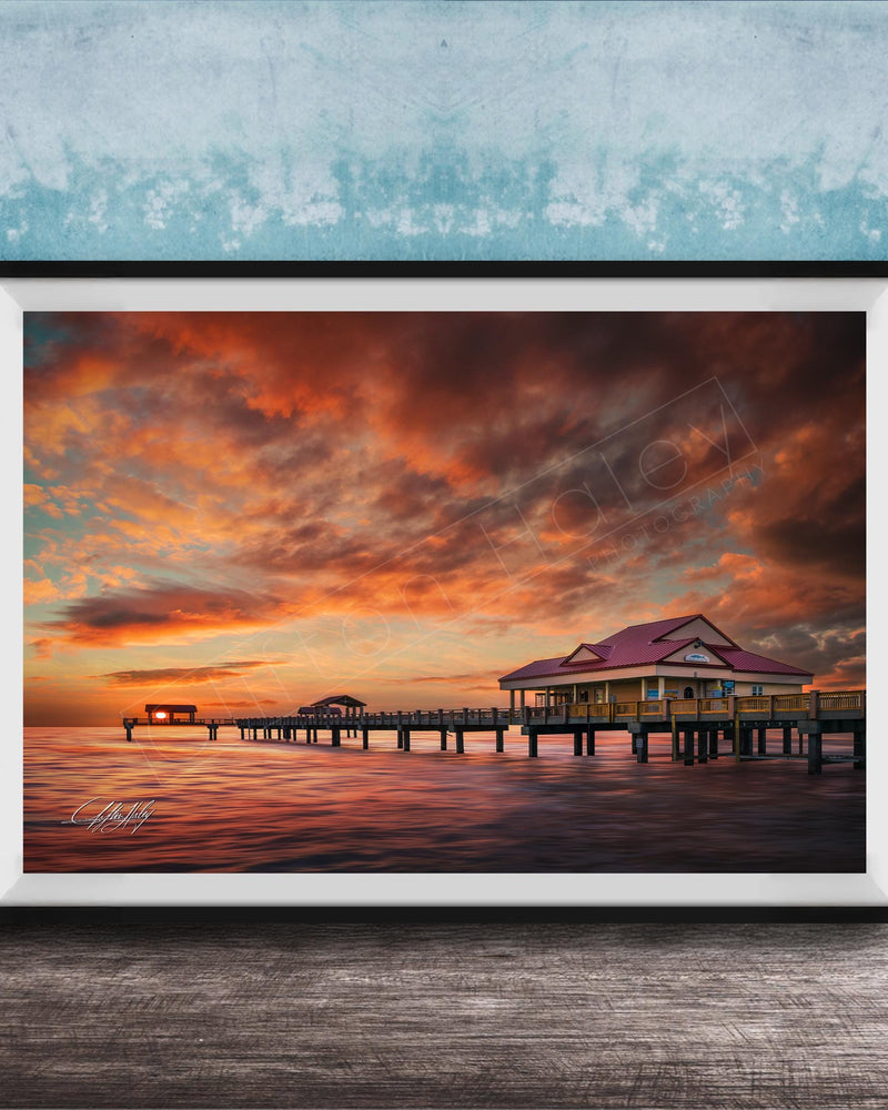 A framed photograph depicting a serene sunset over a wooden pier, with a small building on the pier and a calm body of water in the foreground.