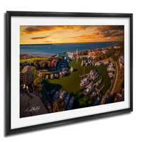 The image is a framed aerial view of a harbor with boats docked at the marina, set against a backdrop of a colorful sunset sky.