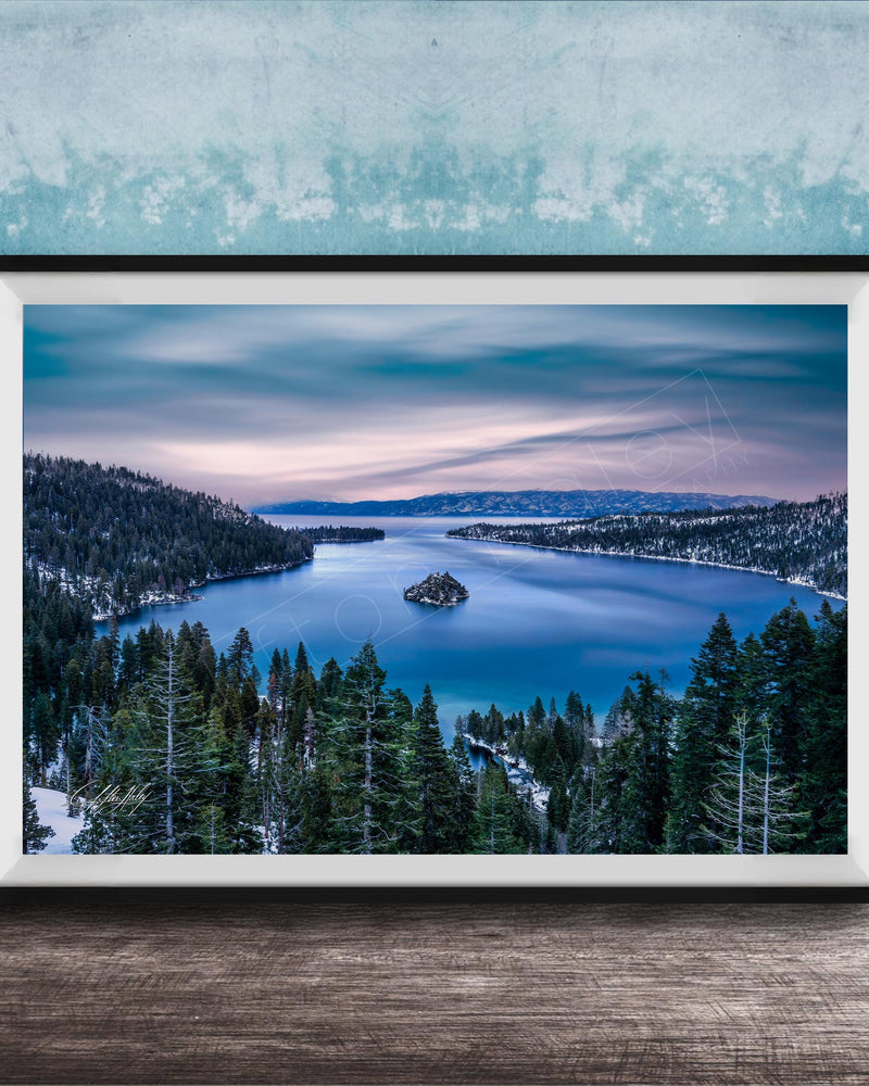 A framed photograph depicting a serene landscape with a lake surrounded by snow-covered trees and mountains in the distance, set against a blue sky with wispy clouds.