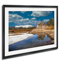 A picturesque landscape with a stone building nestled among snow-capped mountains, surrounded by a serene lake and a clear blue sky with fluffy white clouds.
