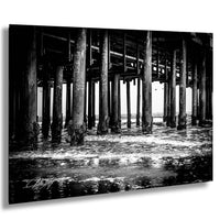 A black and white photograph of a pier with multiple wooden columns and beams, with water flowing under the structure.