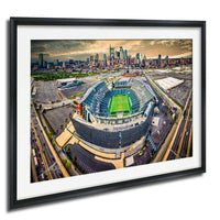 An aerial view of a large sports stadium surrounded by a city skyline, with the stadium's field and surrounding parking lots visible.
