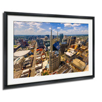 The image is a framed aerial view of a bustling city, showcasing a variety of tall buildings, skyscrapers, and other structures. The cityscape is vibrant and densely populated, with a mix of architectural styles and colors. The image is framed within a black border, and the overall composition is visually striking and dynamic.