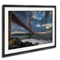 a large suspension bridge spanning a body of water, with dramatic clouds and a sunset sky in the background.