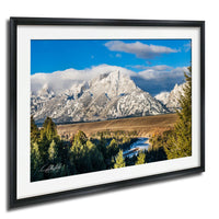 A framed photograph depicting a scenic mountain landscape with snow-capped peaks, a winding river, and a mix of evergreen and deciduous trees.