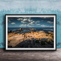 The image is a framed photograph of a rocky beach with a body of water in the background, set against a blue sky with clouds.