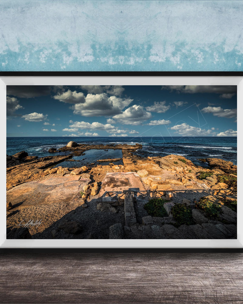 The image is a framed photograph of a rocky beach with a body of water in the background, set against a blue sky with clouds.