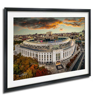 The image is a framed photograph of a large stadium, likely a sports stadium, with a city skyline in the background. The photograph is taken from an aerial perspective, showcasing the stadium's impressive architecture and surrounding urban landscape.