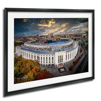 The image is a framed photograph of the Yankee Stadium, a large circular stadium located in New York City. The stadium is surrounded by a cityscape with buildings and trees, and the sky is filled with clouds. The photograph is framed in a black frame and is hung on a wall.