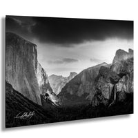 A dramatic black and white landscape photograph depicting a mountainous, forested valley with towering cliffs and a waterfall cascading down the side.