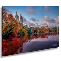 a serene cityscape with a calm lake reflecting the vibrant colors of autumn foliage. The tall buildings in the background are bathed in warm hues, creating a picturesque scene. The sky above is a clear blue, dotted with fluffy white clouds, adding to the overall beauty of the image. The composition of the photograph, with the lake and the buildings in the foreground and the sky in the background, creates a sense of depth and perspective.