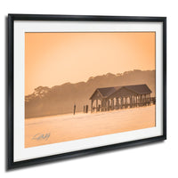 A framed photograph depicting a wooden pier or dock extending into a body of water, with a hazy orange sky in the background.
