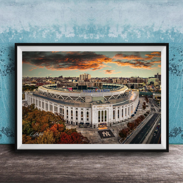 The image is a framed photograph of the New York Yankees stadium, which is a large, circular stadium located in the city of New York. The photograph captures the stadium's distinctive architecture and surrounding cityscape, with a dramatic sky in the background.