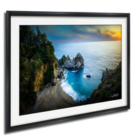 The image is a framed photograph depicting a scenic coastal landscape with a rocky cliff overlooking a beach and the ocean.