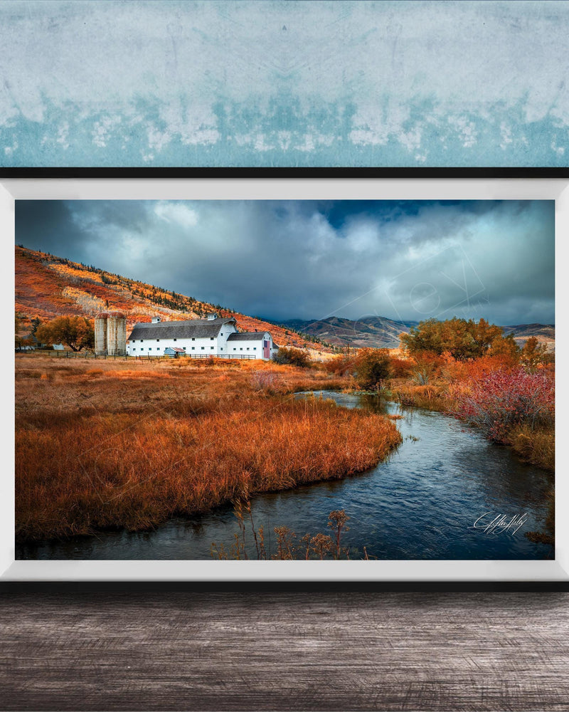 a picturesque rural landscape with a small white building nestled among the rolling hills and lush greenery. The scene is framed and displayed on a wooden surface, with a blue wall in the background.