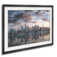 a city skyline at sunset, with the tall buildings reflected in the calm water below.