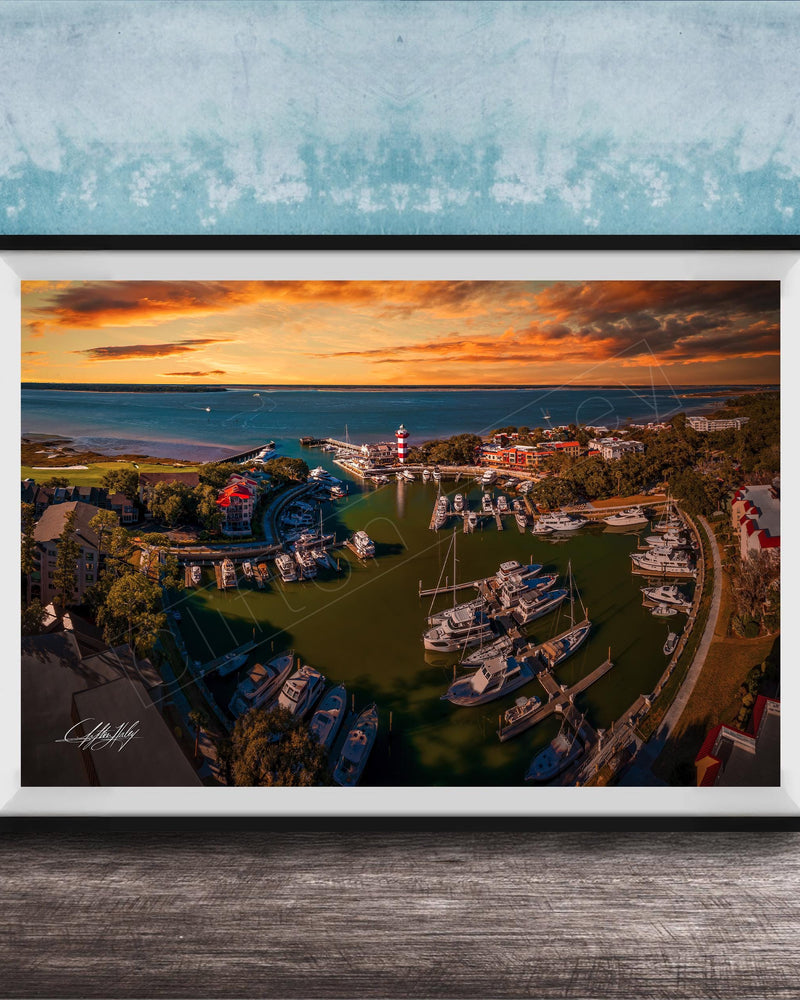 The image is a framed aerial view of a harbor with boats docked, set against a backdrop of a colorful sunset sky.