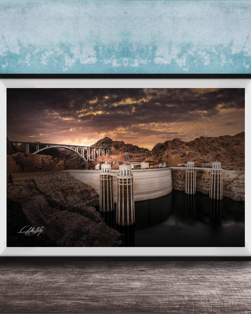 A framed photograph depicting a large dam with a bridge spanning across it, set against a dramatic sky with clouds and a sunset.