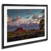 A rustic wooden barn sits in a grassy field, with majestic mountains rising in the background under a vibrant sky.