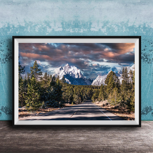 A framed photograph depicting a scenic mountain landscape with a winding road surrounded by pine trees and snow-capped peaks under a dramatic cloudy sky.