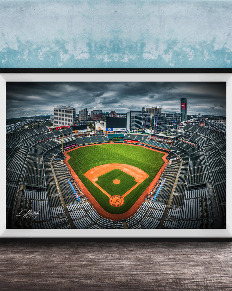 A framed photograph of a baseball stadium with a large field, surrounded by buildings and a cloudy sky.