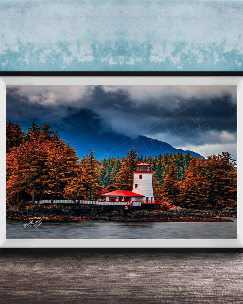 A framed photograph depicting a lighthouse surrounded by a forest with autumn-colored trees, set against a cloudy sky.