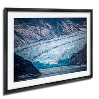 A framed photograph depicting a large, blue-green glacier with ice formations and a body of water in the foreground.