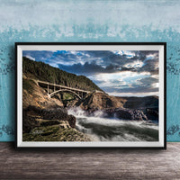 a scenic view of a bridge spanning over a rushing river, with a dramatic sky and lush green trees in the background.