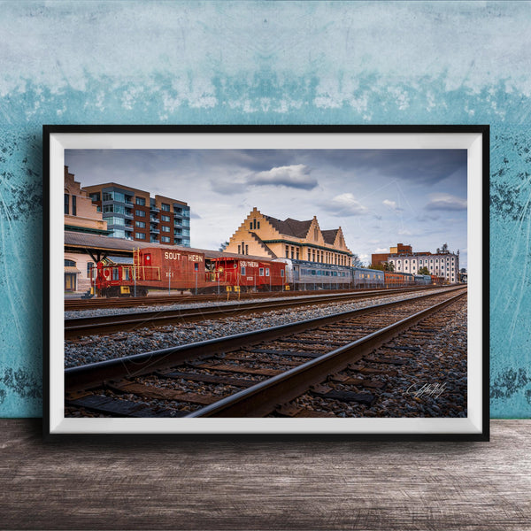 A framed photograph of a train on railroad tracks, with buildings in the background, set against a blue wall.