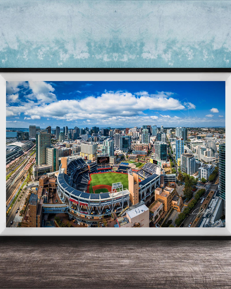 An aerial view of a large city with a baseball stadium in the center, surrounded by tall buildings and a blue sky with clouds.