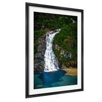 A framed photograph depicting a majestic waterfall cascading down a rocky cliff, surrounded by lush greenery and a serene blue-green body of water.