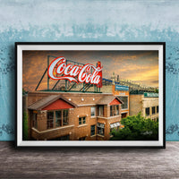 A framed photograph of a Coca-Cola sign on top of a brick building, with a sunset sky in the background.