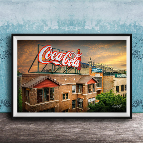 A framed photograph of a Coca-Cola sign on top of a brick building, with a sunset sky in the background.
