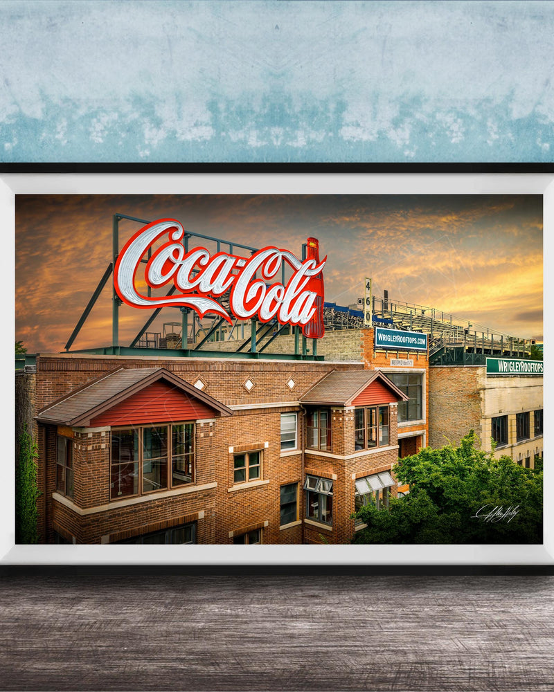 A framed photograph of a Coca-Cola sign on top of a brick building, with a sunset sky in the background.