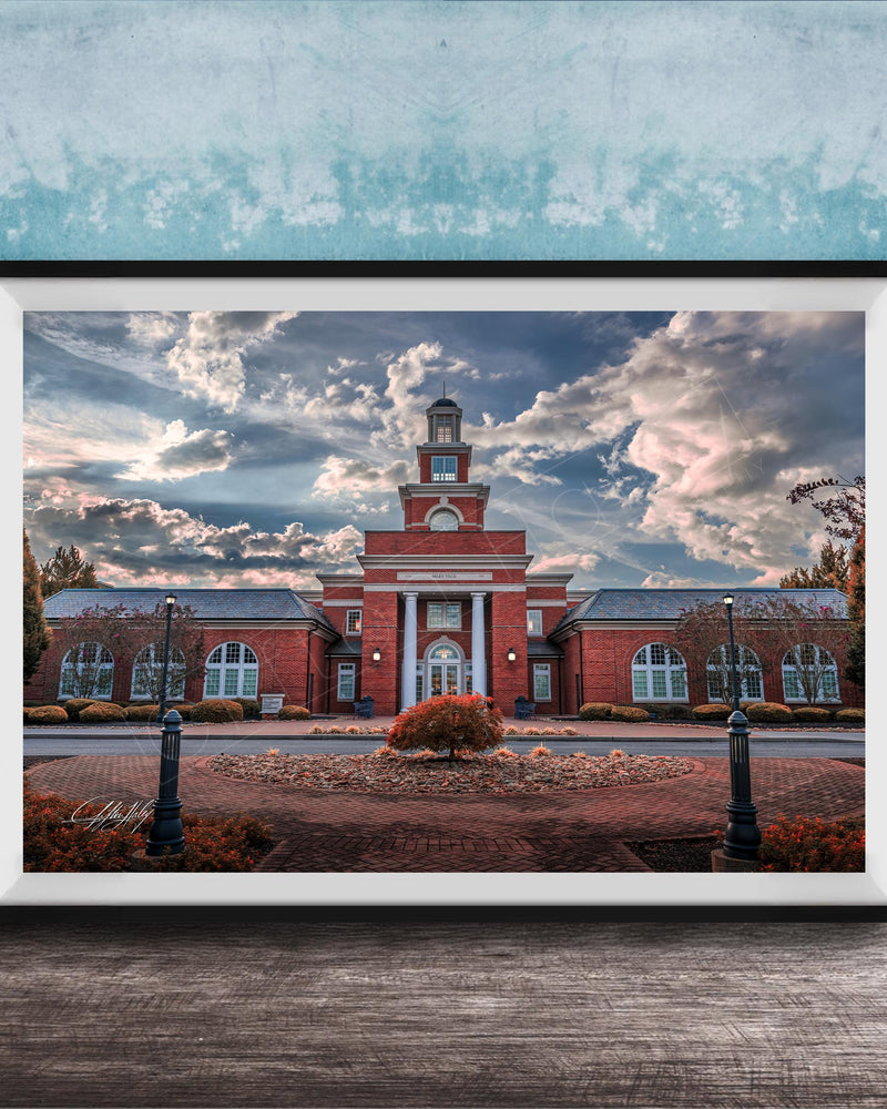 A framed photograph of a red brick building with a clock tower, surrounded by a brick walkway and landscaping, set against a cloudy sky.