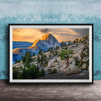 A framed photograph depicting a mountainous landscape with a large, snow-capped peak in the distance, surrounded by trees and rocky terrain. The sky is filled with warm, golden light, creating a serene and picturesque scene.