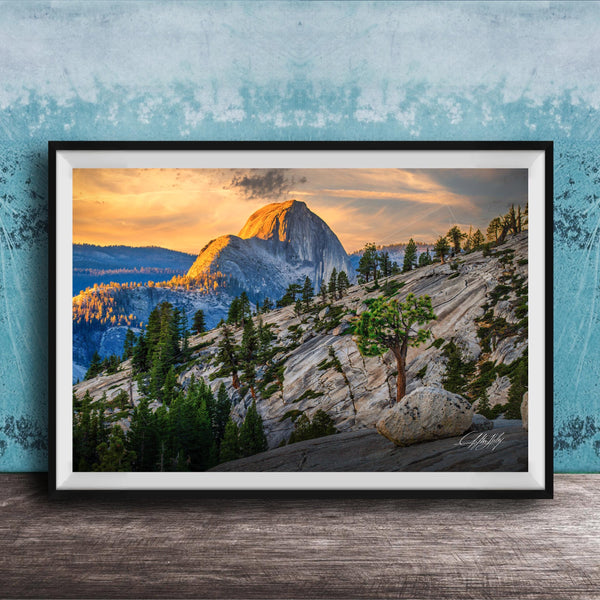 A framed photograph depicting a mountainous landscape with a large, snow-capped peak in the distance, surrounded by trees and rocky terrain. The sky is filled with warm, golden light, creating a serene and picturesque scene.