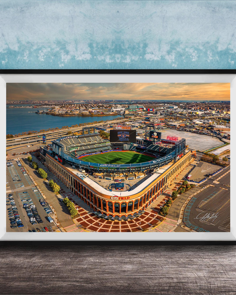 An aerial view of a baseball stadium with a large field, surrounding parking lots, and a body of water in the background.