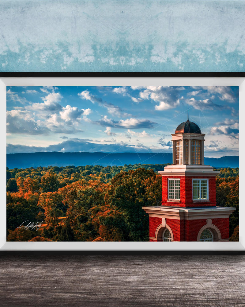 A framed photograph of a red brick building with a dome, set against a backdrop of a blue sky with clouds and a mountainous landscape.