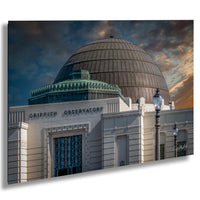 the Griffith Observatory, a large domed building with a distinctive architectural style, set against a dramatic sky with clouds and a sunset.