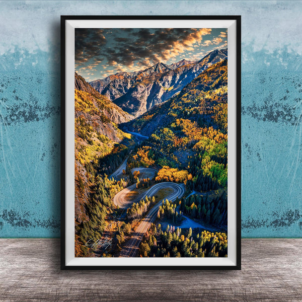 The image is a framed photograph of a winding mountain road surrounded by lush forests and autumnal trees, with a dramatic sky in the background.