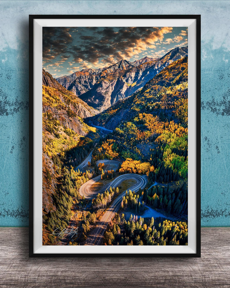 The image is a framed photograph of a winding mountain road surrounded by lush forests and autumnal trees, with a dramatic sky in the background.
